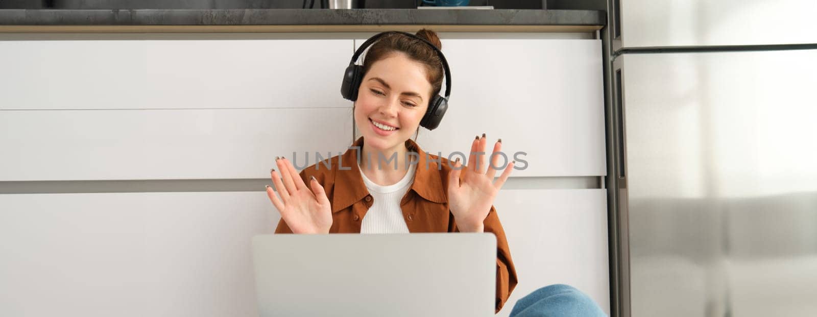 Joyful young woman in headphones, waves hands at laptop, joins online chat, connects to remote course or video call, sits on floor at home in the kitchen.