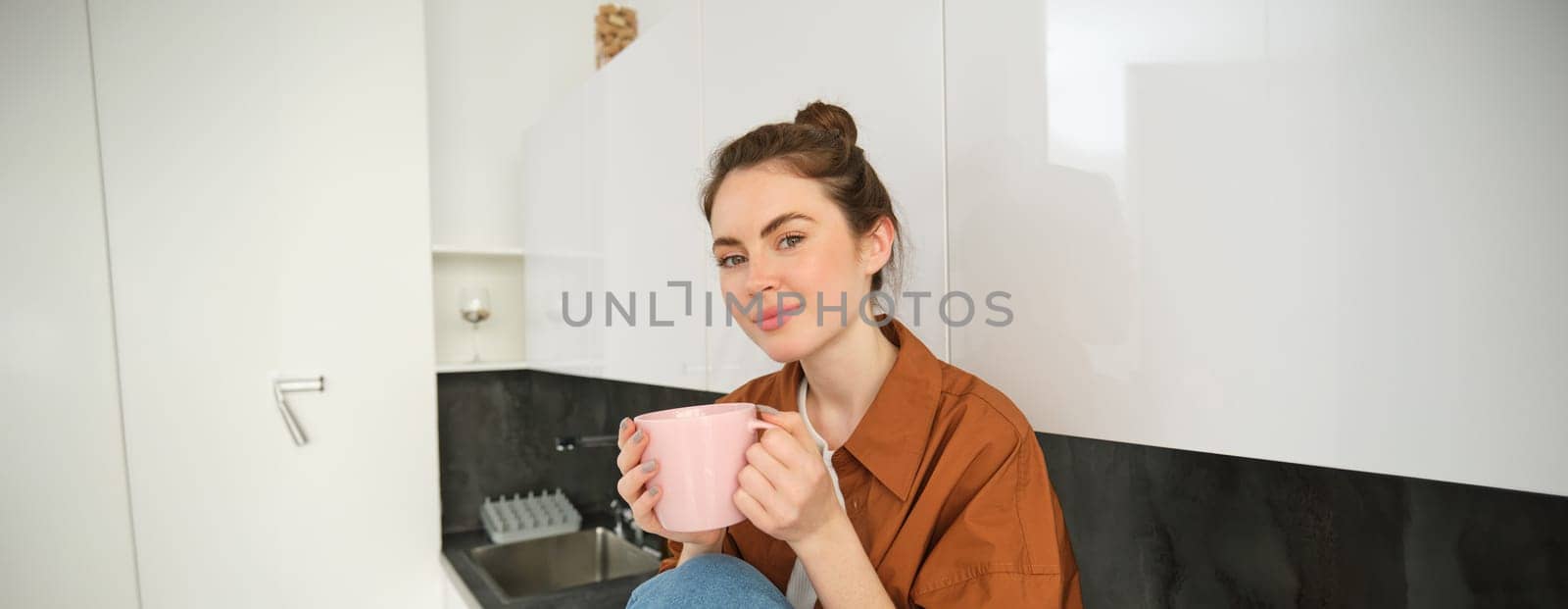 Portrait of beautiful young woman enjoying her morning cup of aromatic coffee, drinking tea while sitting in kitchen and contemplating sunny day.