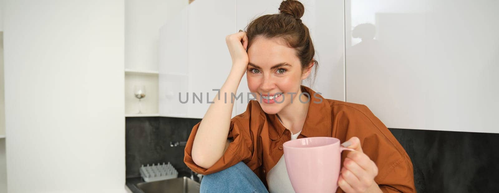 Portrait of young woman relaxing in kitchen with cup of coffee, smiling at camera, sitting on kitchen counter.