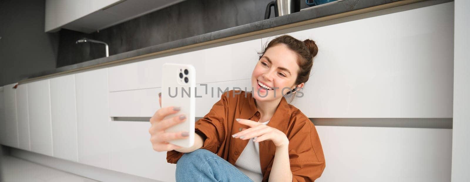 Portrait of beautiful smiling woman, talking to friend on smartphone video chat, connects to online conversation on mobile app, sitting on kitchen floor by Benzoix