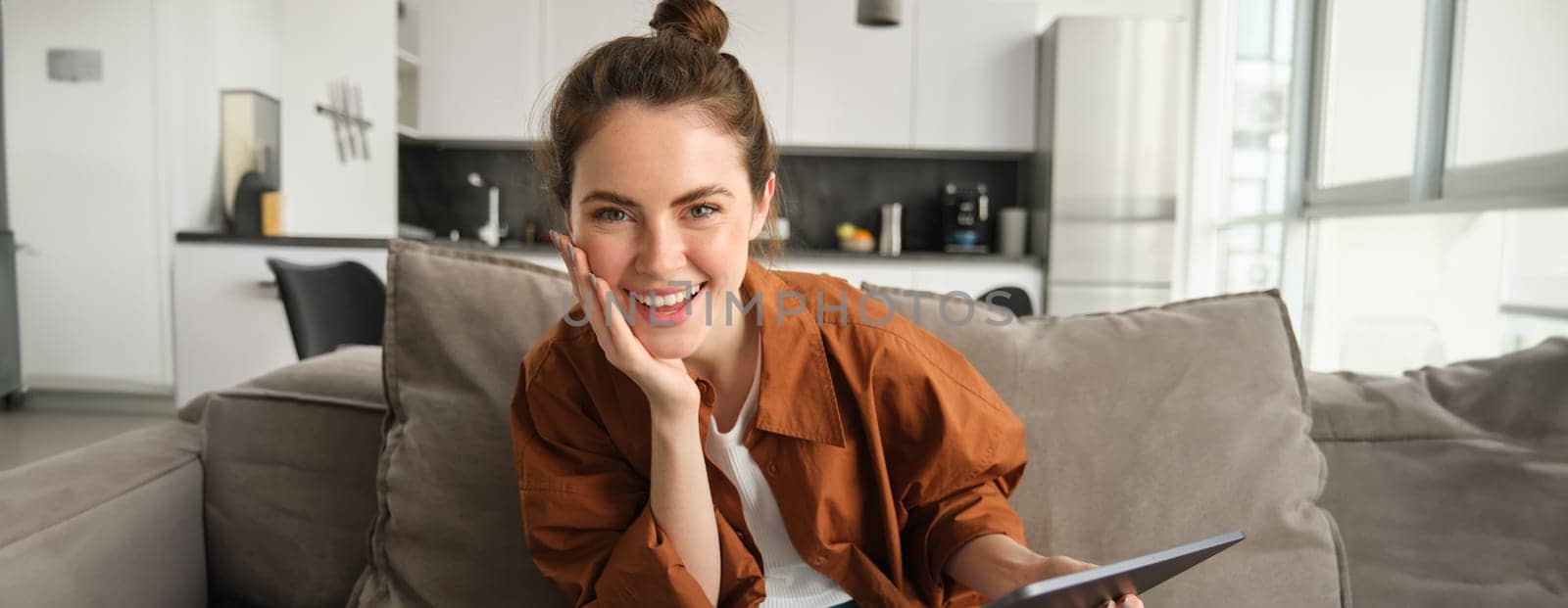 Close up portrait of young modern woman, girl sitting on couch and using digital tablet, reading e-book, laughing and smiling at camera.