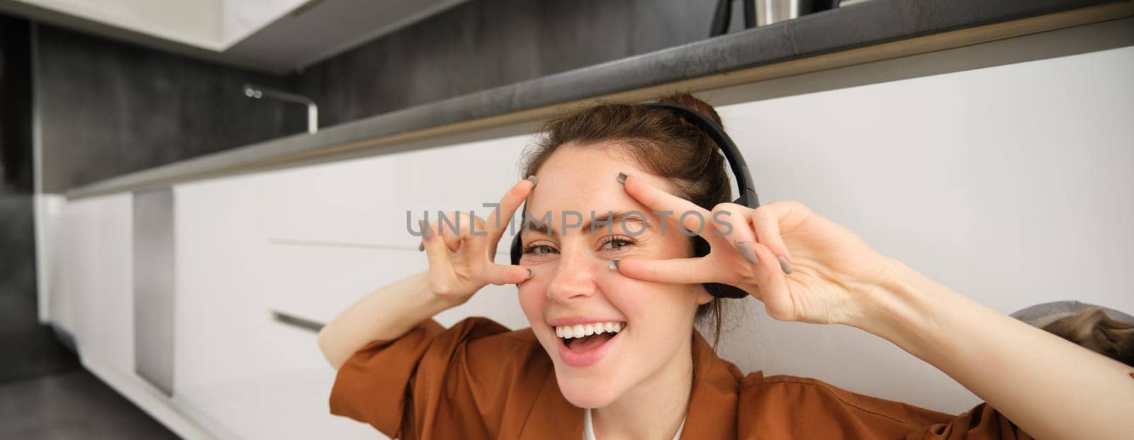 Close up of carefree young woman, laughing and smiling, showing peace, v-sign gesture, listening to music in wireless headphones.