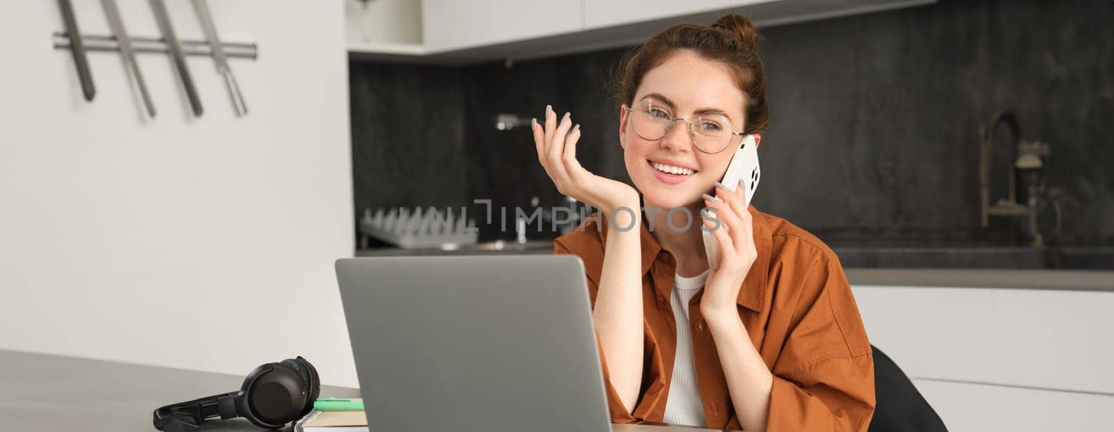 Portrait of beautiful smiling woman working from home, talking on mobile phone, calling client, self-employed businesswoman sets up workplace in kitchen, using laptop and smartphone.