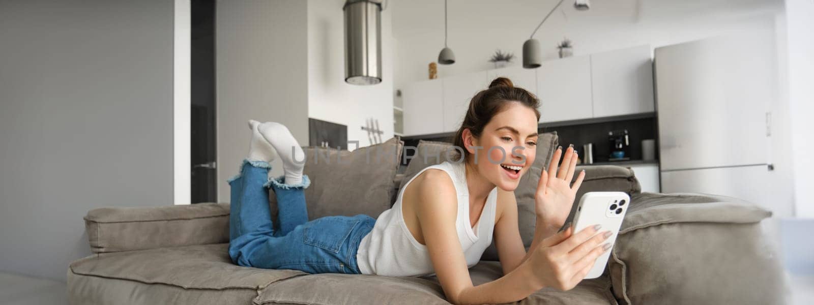 Carefree young smiling woman, lying on sofa, saying hello and waving at smartphone screen, video calling someone, chatting online using mobile app.