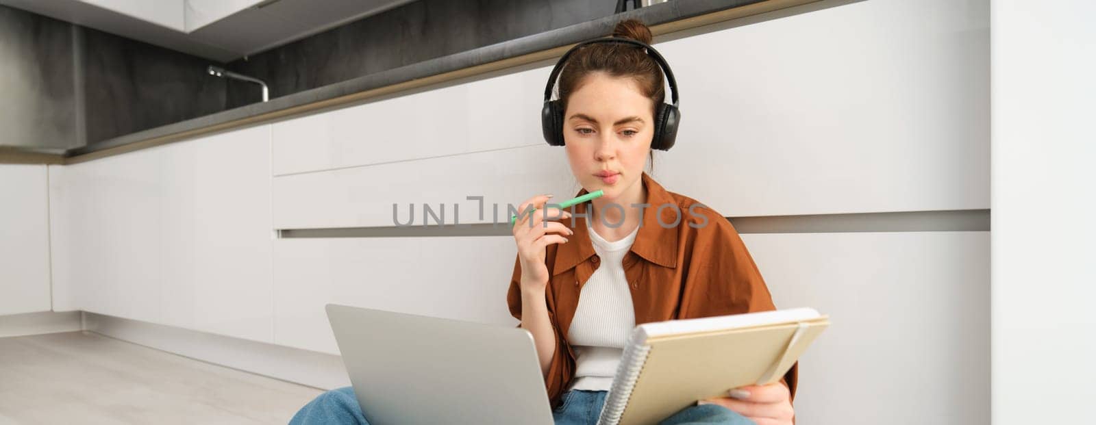 Serious-looking girl student studying at home, sits with laptop and notebook on floor, doing homework project on computer, has online course lesson, wears headphones.