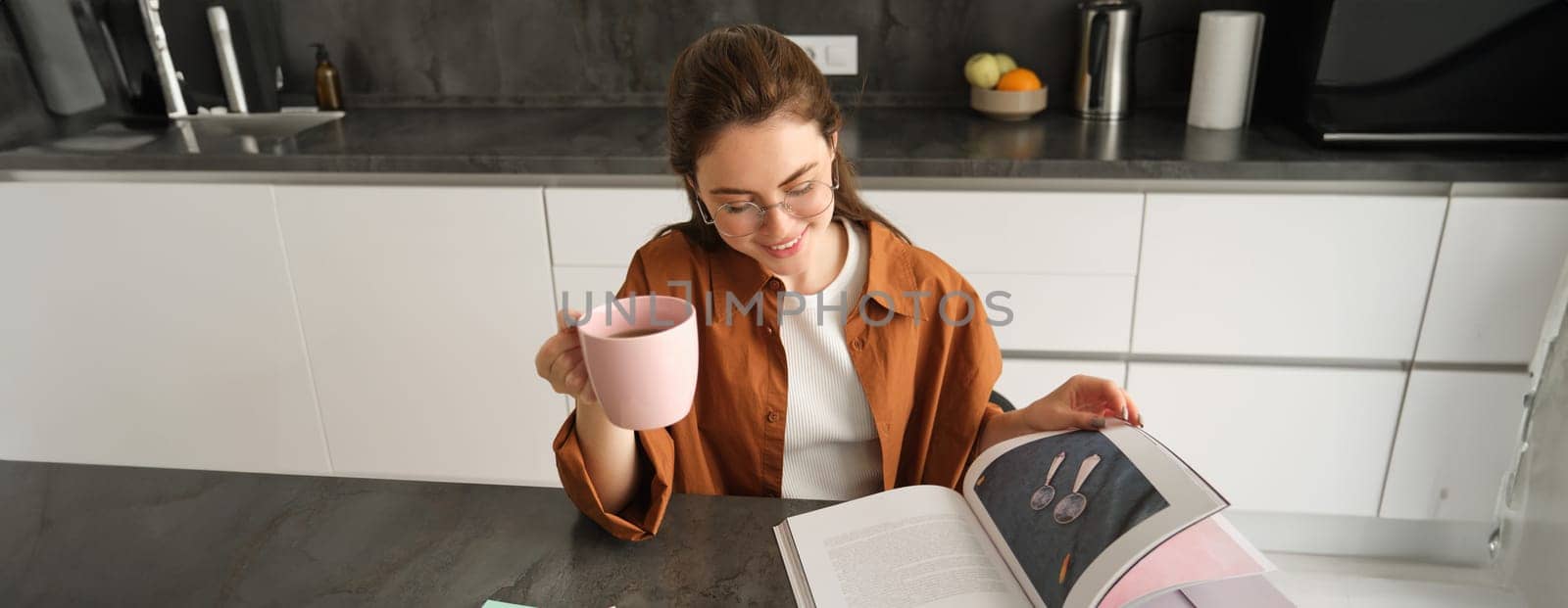 Portrait of beautiful young woman in kitchen, sitting with a book, flipping pages, reading and drinking coffee by Benzoix