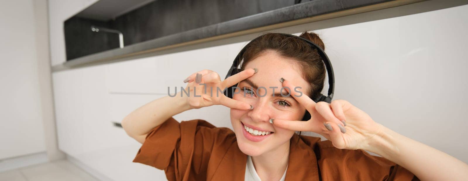 Close up of carefree young modern woman, relaxes at home, listens music in wireless headphones, shows v-sign and smiles at camera.