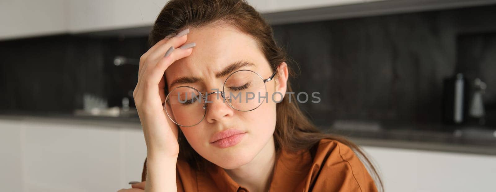 Portrait of tired young working woman in glasses, holds hands on head, suffers painful migraine, has pain in temples, sitting with headache in kitchen by Benzoix