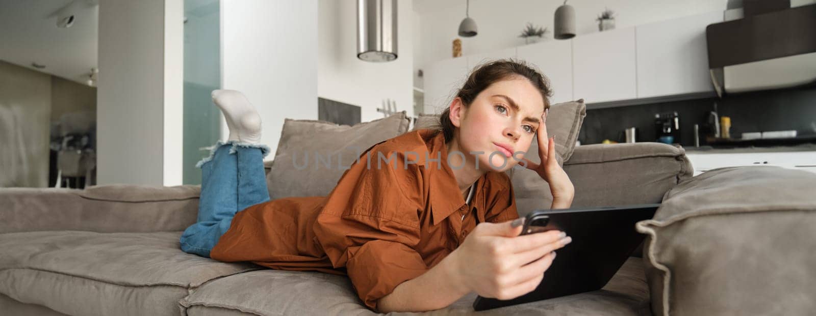 Portrait of woman lying on couch at home, holding digital tablet and looking thoughtful, frowning while thinking and looking aside, making decision, purchase online on gadget by Benzoix