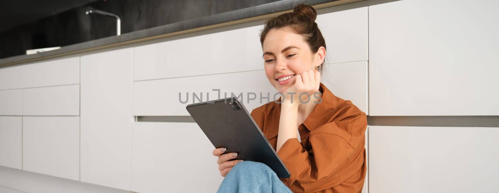 Portrait of beautiful, carefree young woman, sitting on kitchen floor with digital tablet, working remote from home, using device to check her mail. Girl watching something on her device.