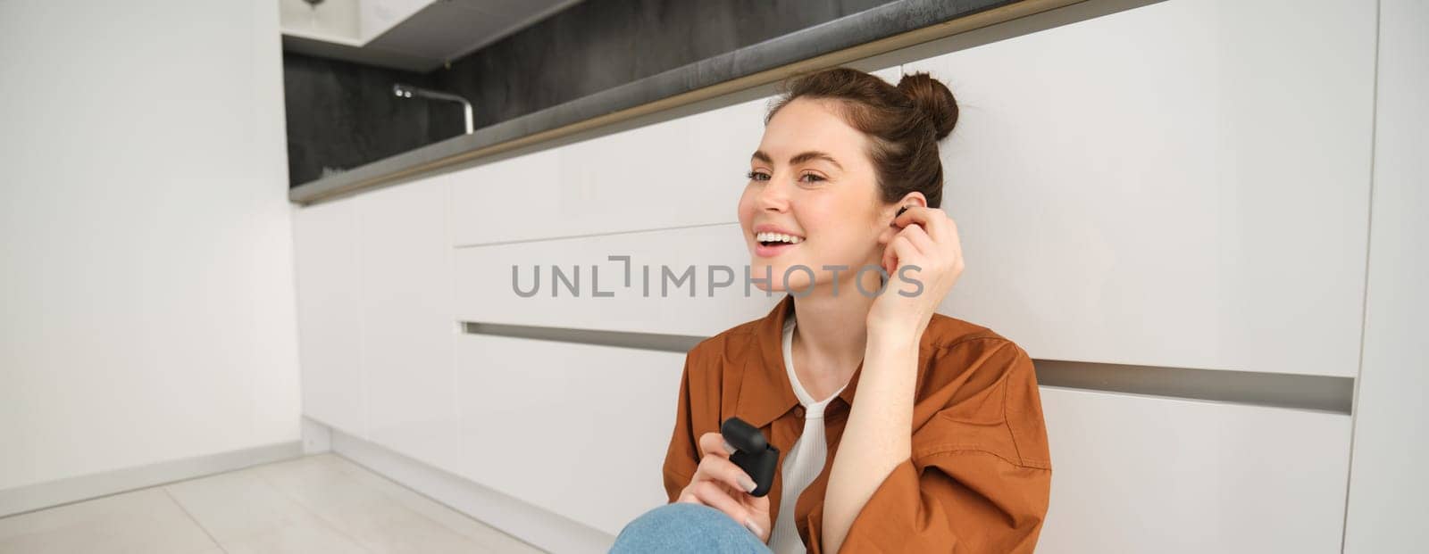 Portrait of young smiling woman, girl puts on her black wireless headphones, listens to music in earphones, sits on kitchen floor at home.