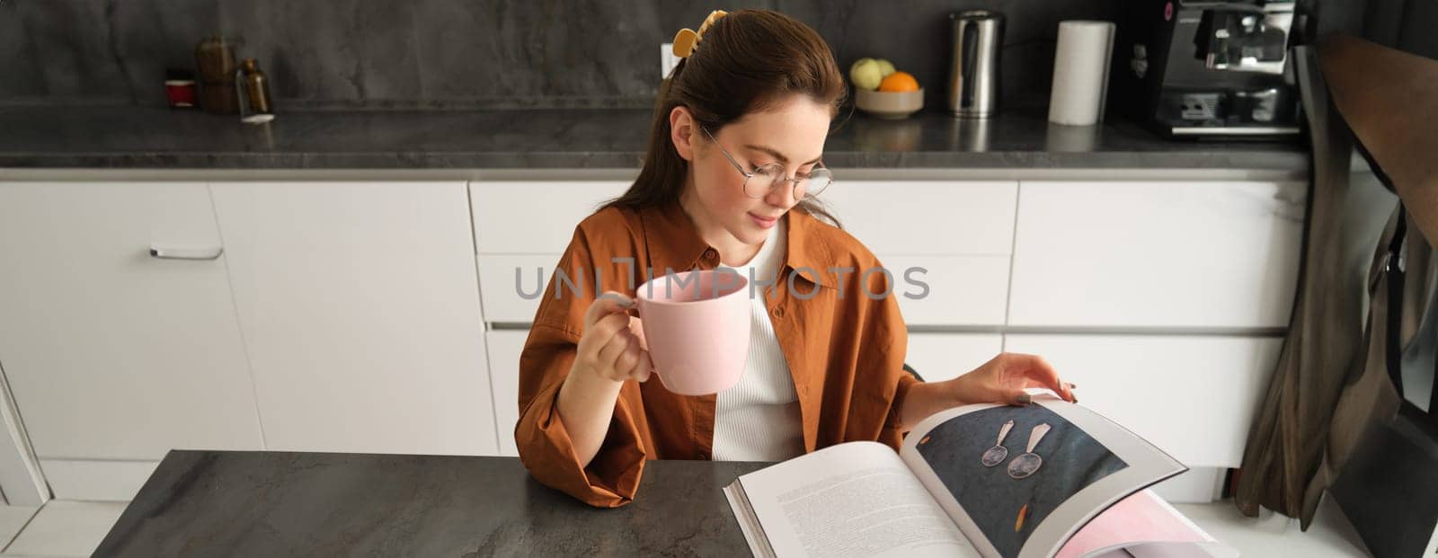 Portrait of beautiful young woman in kitchen, sitting with a book, flipping pages, reading and drinking coffee by Benzoix