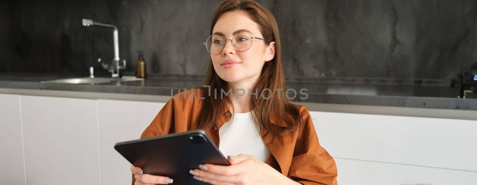 Image of beautiful young woman in glasses, sitting in her kitchen with digital tablet, watching videos online, reading, working on remote, studying remotely by Benzoix