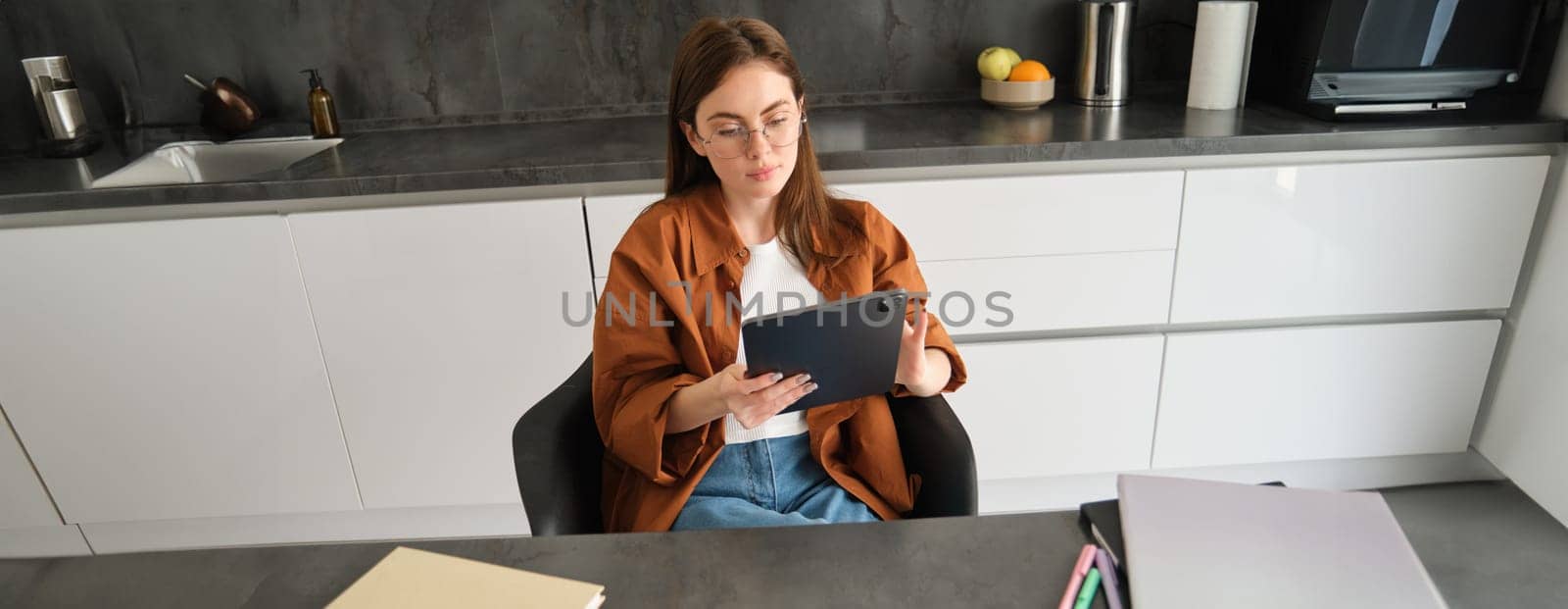 E-learning and remote education concept. Young woman in glasses, works from home in kitchen, looking at digital tablet, connects to online class or course.