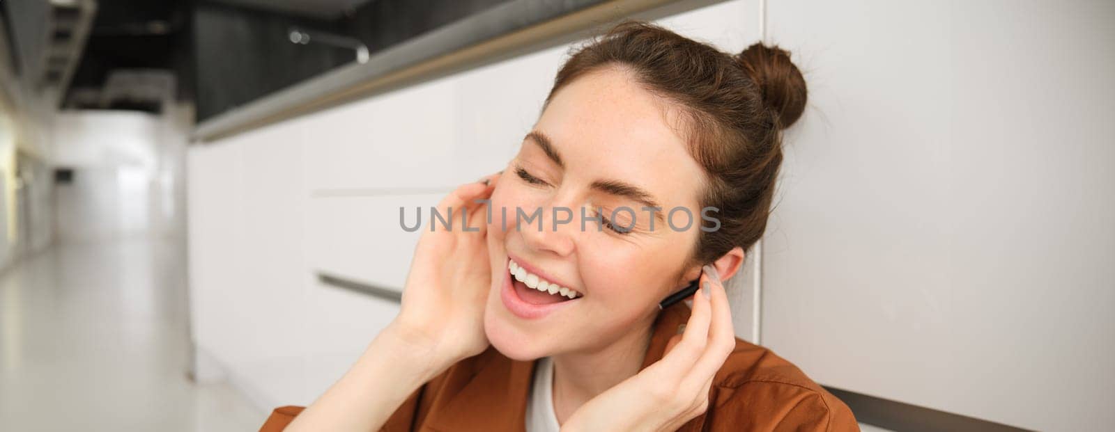 Close up portrait of girl enjoying listening to music in wireless earphones, smiling, wearing headphones, sitting at home by Benzoix