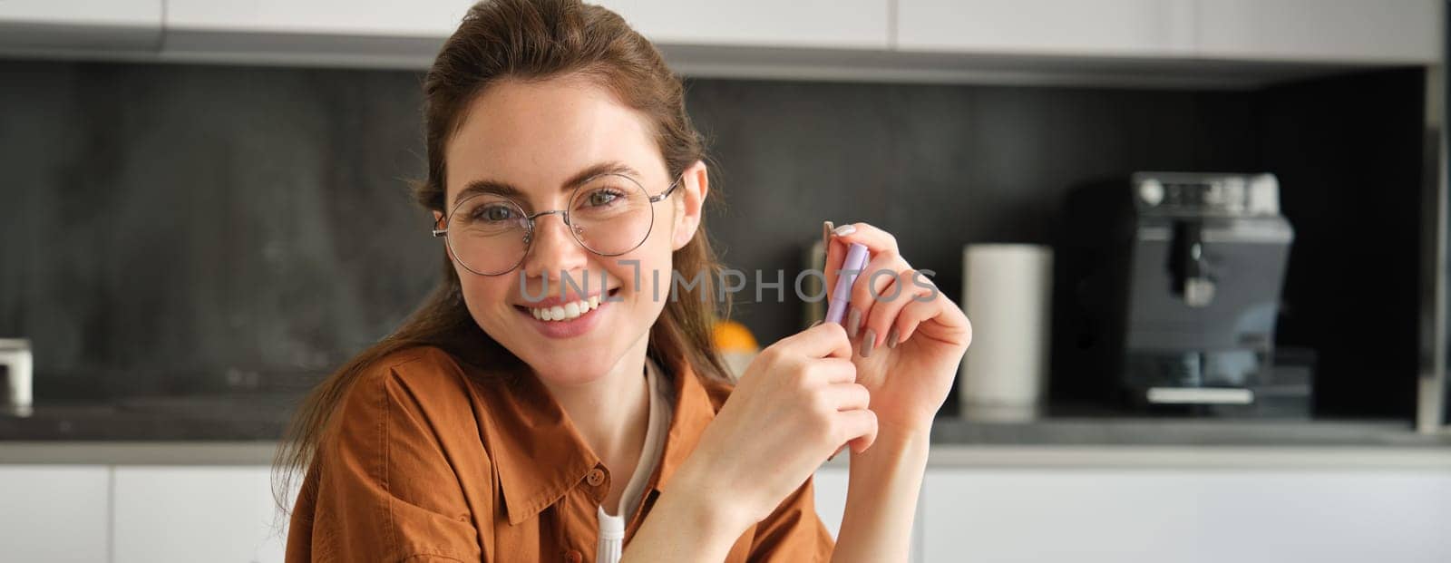 Portrait of beautiful young woman, entrepreneur in glasses, has own business from home, sitting in kitchen with pen, smiling and looking confident at camera by Benzoix