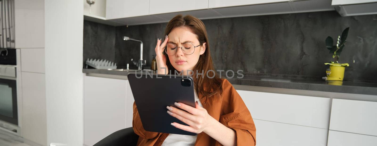 Portrait of woman freelancer, wearing glasses, holding digital tablet, looking tired, has migraine or headache, reading e-book. Freelance worker sits at home with gadget.