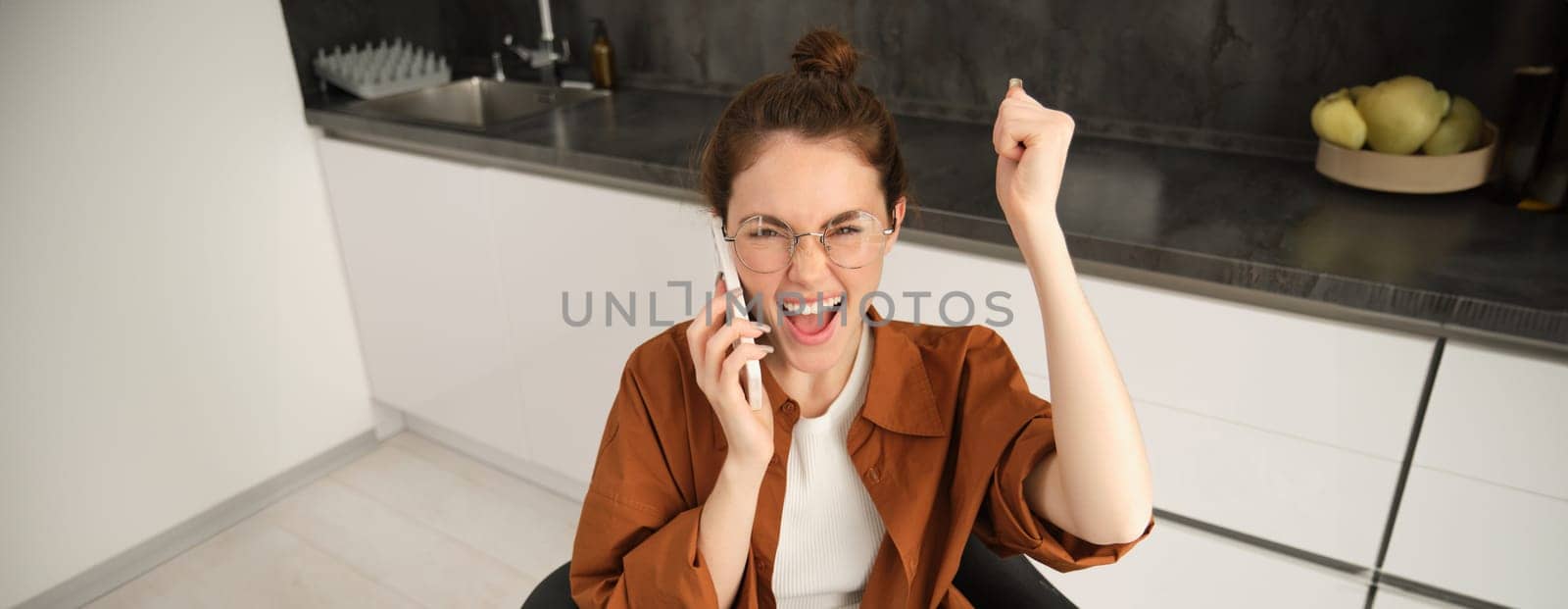 Portrait of excited woman in kitchen, sitting with mobile phone, celebrating, receives great news over the telephone, triumphing by Benzoix