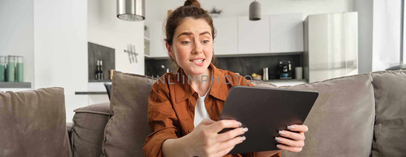 Portrait of young woman looking stressed and tensed at digital tablet, playing difficult level video game, sitting on couch, using gadget, spending time at home.