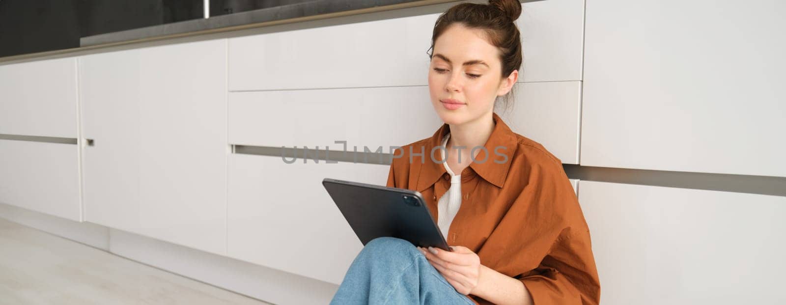 Portrait of cute young woman, freelancer working from home, sitting on kitchen floor with digital tablet and smiling.