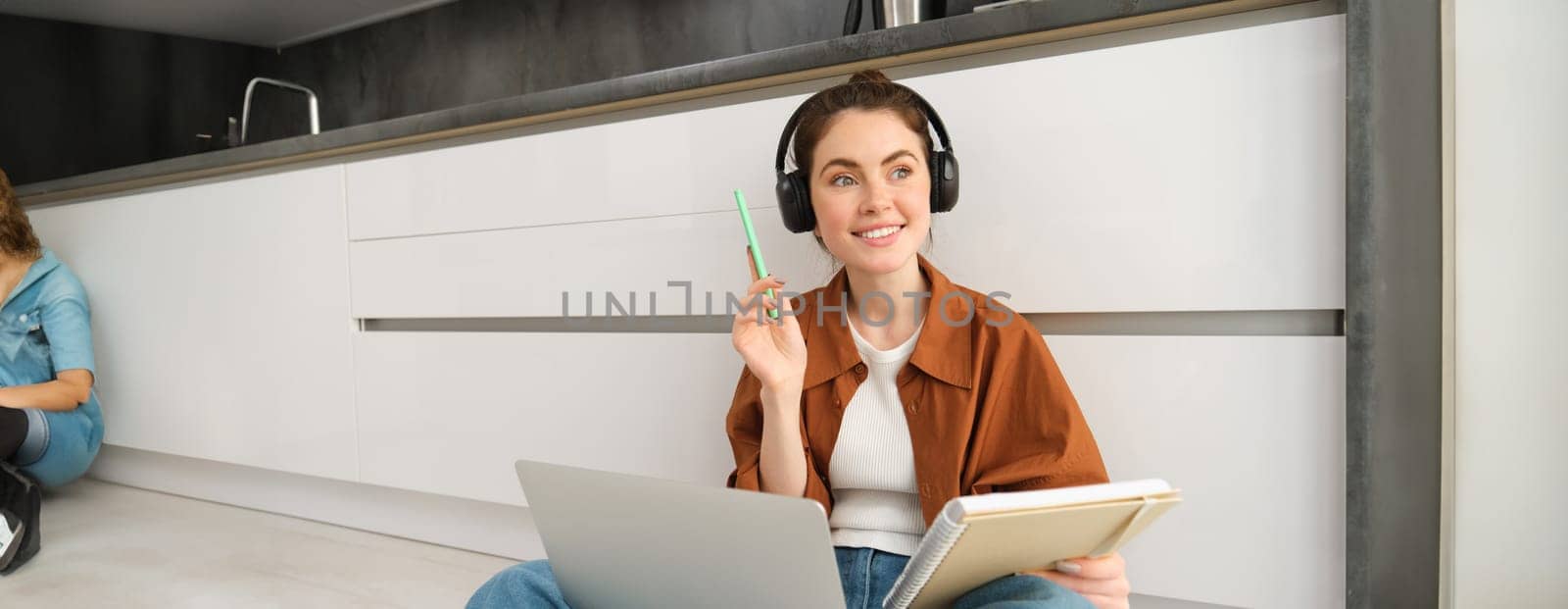 Enthusiastic young woman, found a solution and looks happy, sits on floor and does homework on laptop, makes notes in notebook, holds pen, studies in wireless headphones.