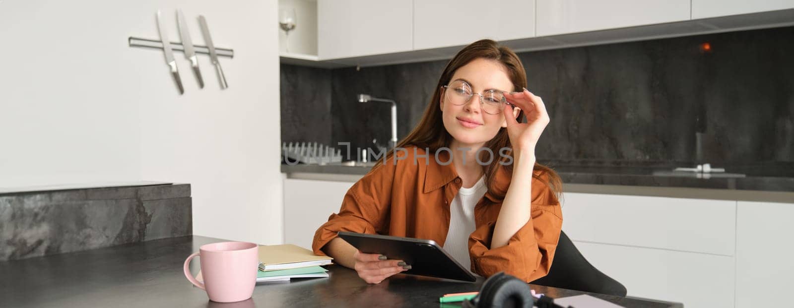 Portrait of young student, woman studying at home, working remotely, setup workplace in her kitchen, sitting on chair with digital tablet, reading in glasses by Benzoix