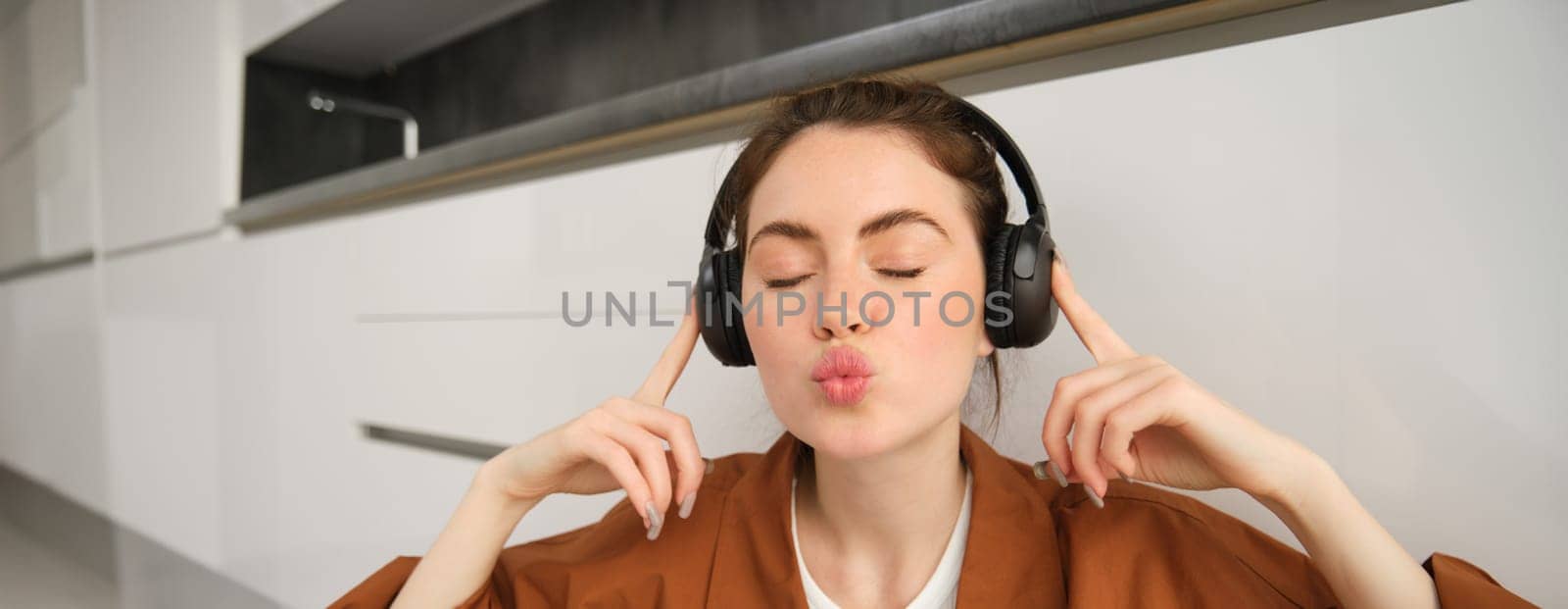 Close up of cute young woman, listens music in wireless headphones, makes kissing puckered lips and closes eyes, poses in earphones by Benzoix