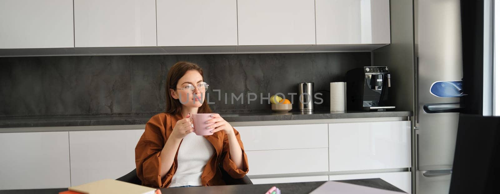 Portrait of young woman enjoying cup of coffee in peace, sitting at home, holding mug with herbal tea, wearing glasses, relaxing at in kitchen by Benzoix