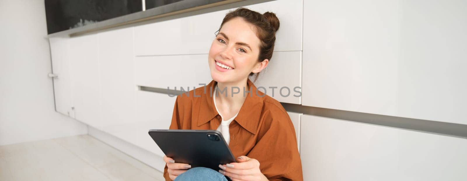 Young entrepreneurs. Smiling cute woman with digital tablet, sitting with gadget on kitchen floor and looking happy, studying remotely, reading on her device by Benzoix