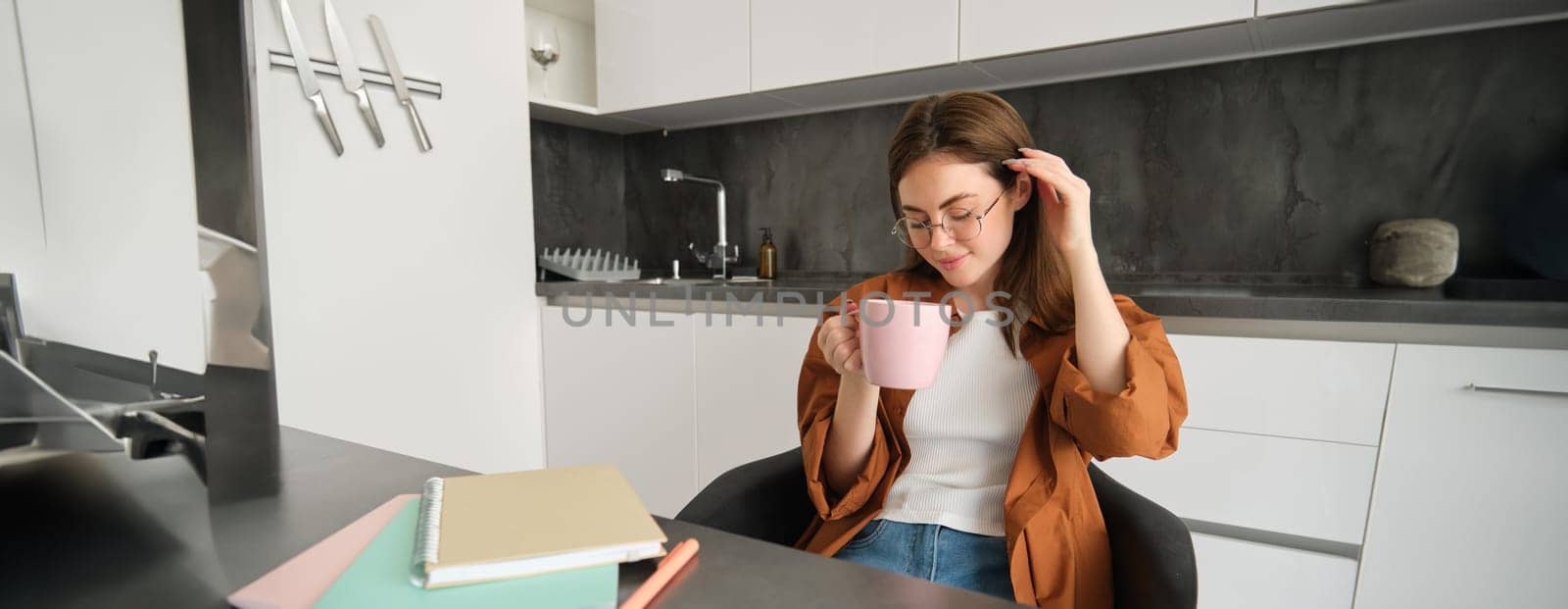 Portrait of young woman working from home, taking a break for cup of tea. Girl studies remotely, sits in kitchen, drinking hot coffee and smiles by Benzoix