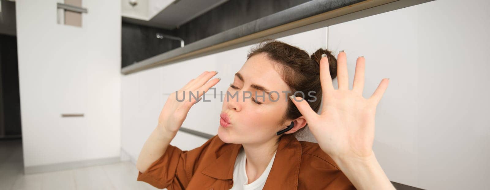 Dancing girl sits on kitchen floor, moving to the rhythm, enjoying sound quality in wireless headphones, listening to music in earphones by Benzoix