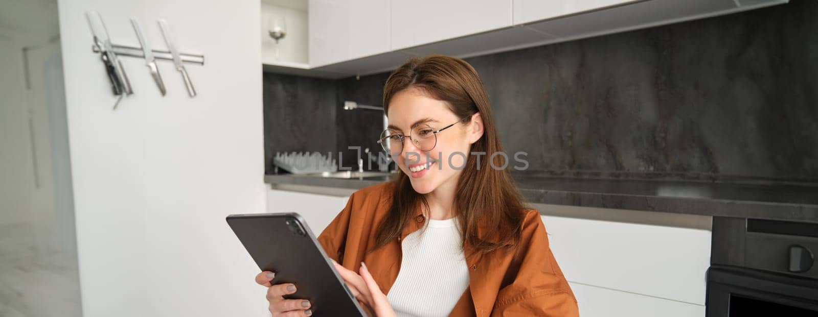 Portrait of girl in glasses, using digital tablet in kitchen, working from home, touches screen and smiling by Benzoix