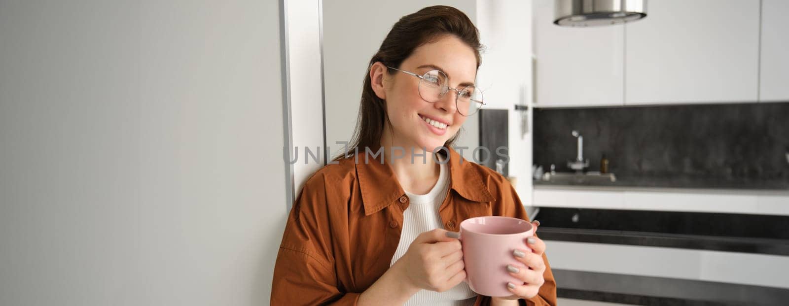 Portrait of beautiful, smiling female model, woman in glasses with cup of coffee, leans on wall at home, drinks tea.