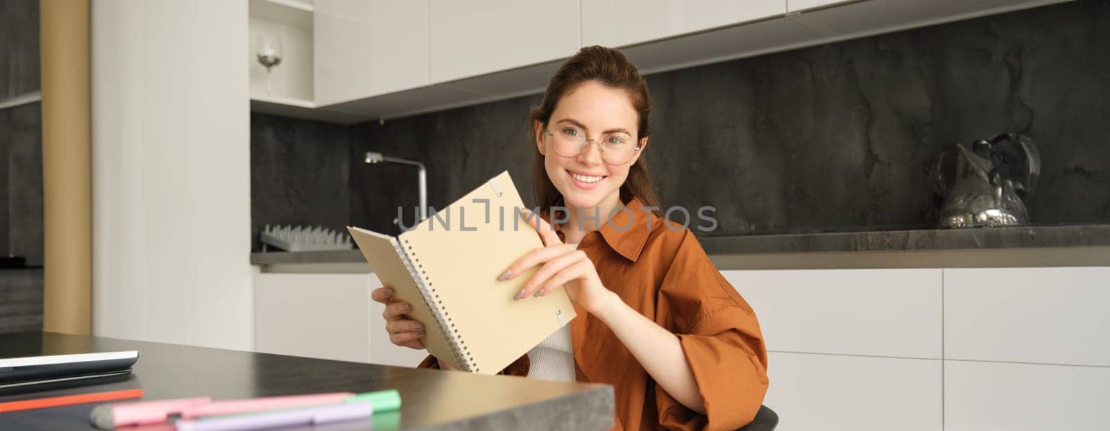 Close up portrait of young woman, student in kitchen, holding notebook, revising for exam at home, studying, reading her planner.