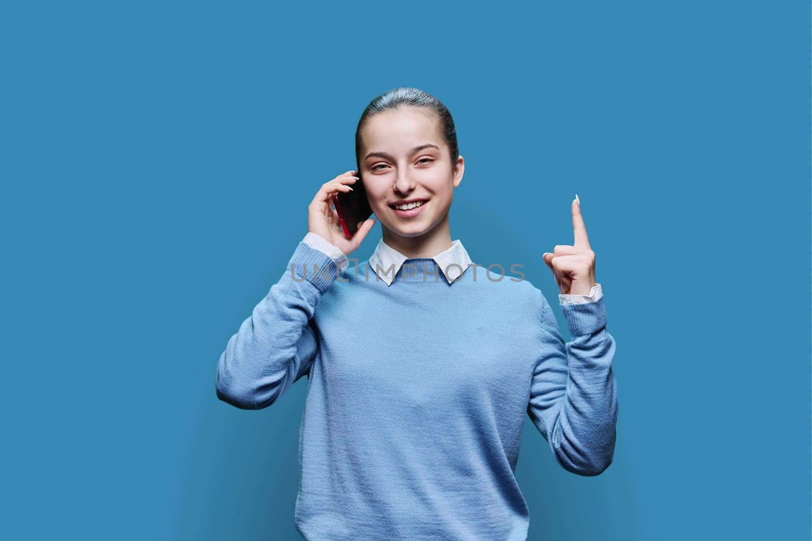 Joyful emotional surprised happy cheerful teenage student, girl 15, 16 years old talking on mobile phone on blue studio background. Lifestyle, communication, adolescence concept