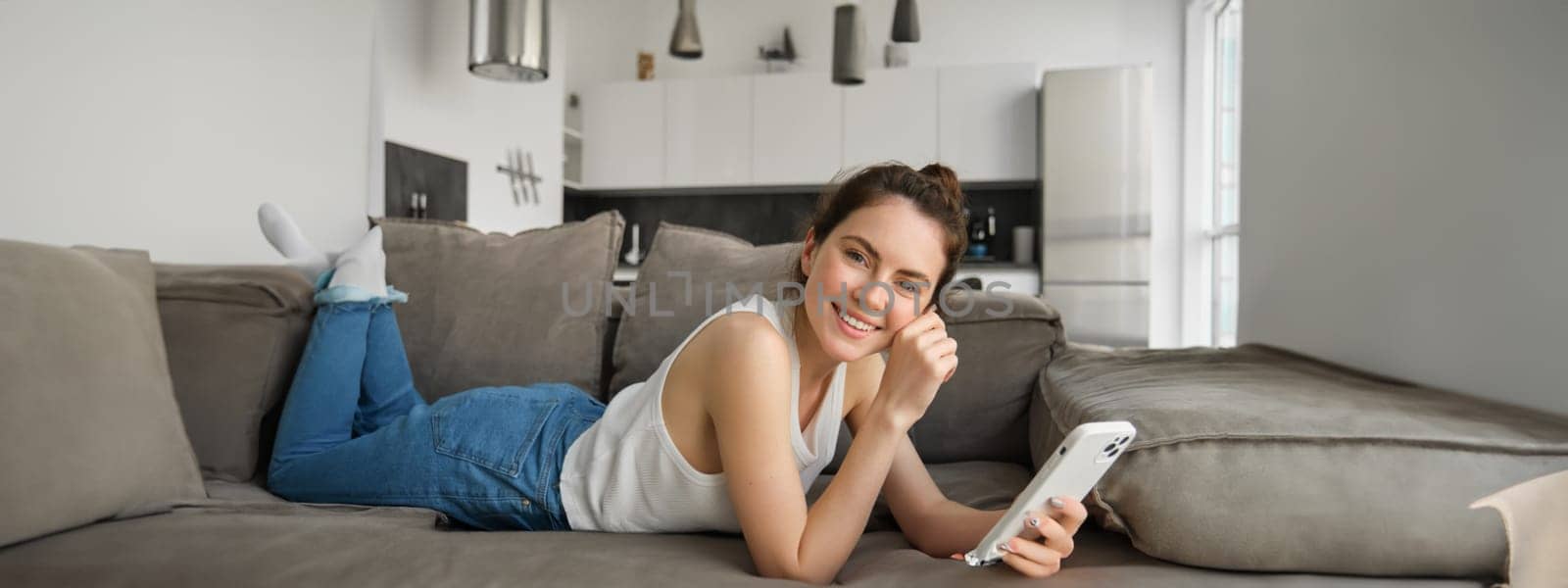 Portrait of cute, happy young woman lying on couch, holding mobile phone, using smartphone while relaxing at home in living room, concept of lifestyle and people leisure.