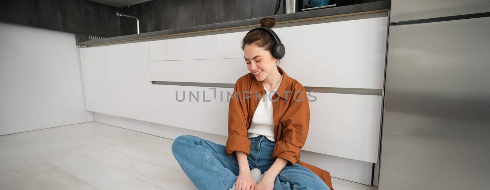 Cute modern girl on kitchen floor at home, listening music in wireless headphones, having fun.
