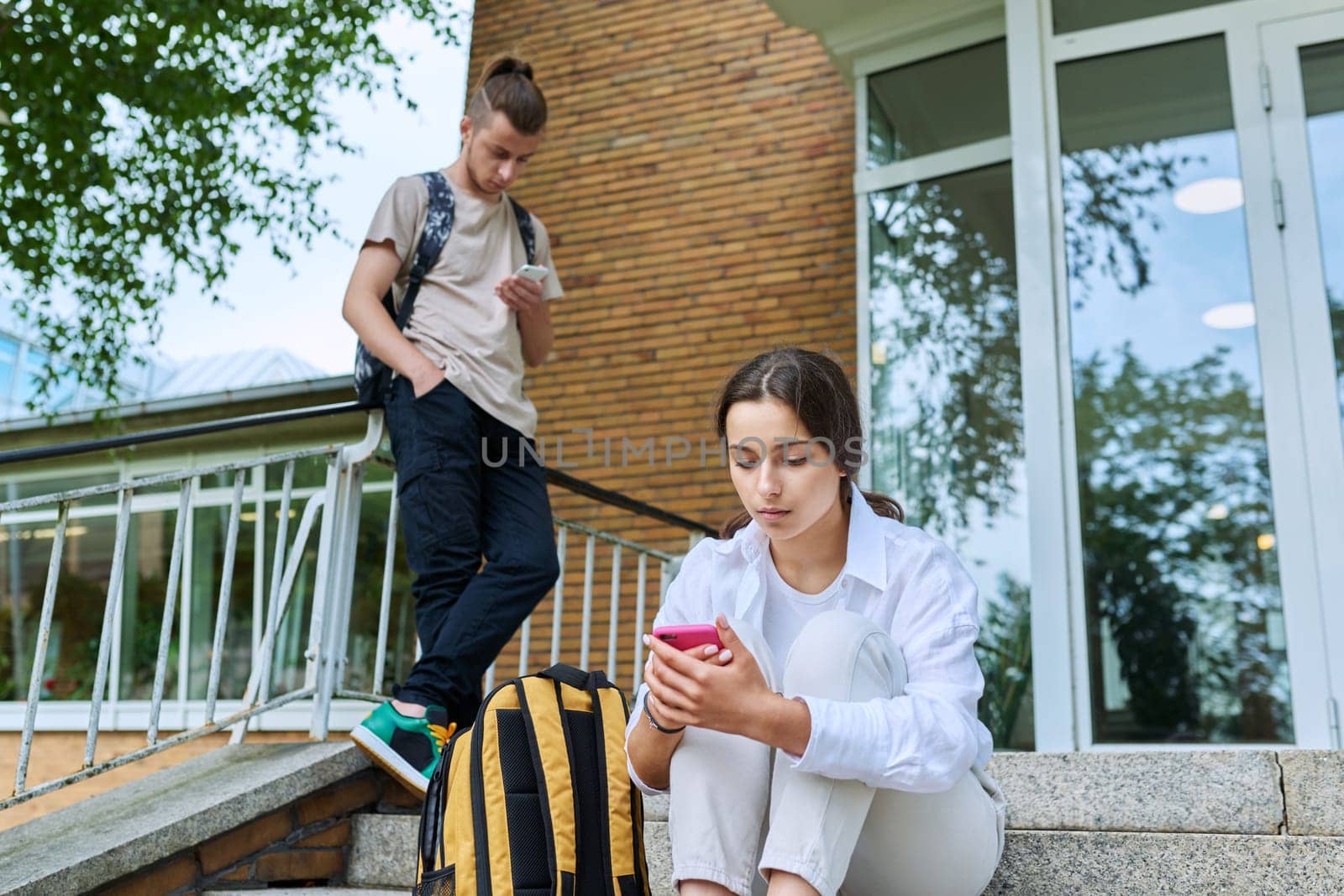 Teenage students with smartphones on the steps of educational building, in focus teenager girl high school age using gadget. Adolescence, youth, education, lifestyle, technology concept