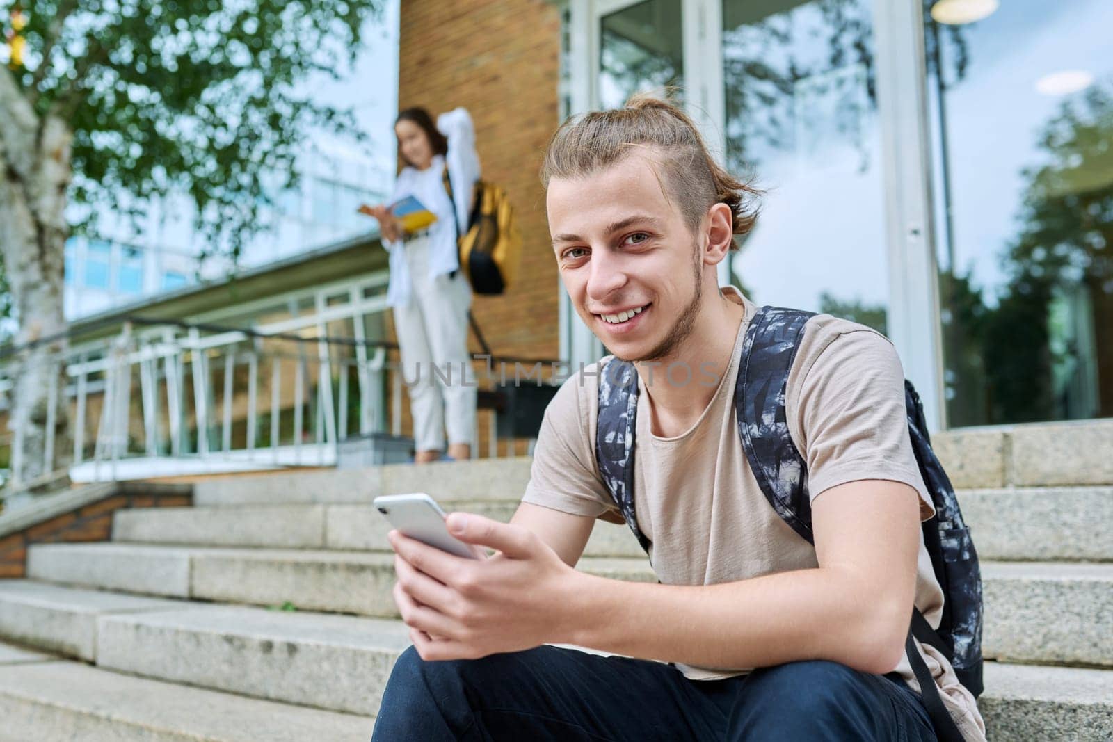 Hipster guy smiling teenager student 18, 19 years old with backpack smartphone looking at camera, sitting on steps of outdoor educational building. Youth, education, lifestyle, technology