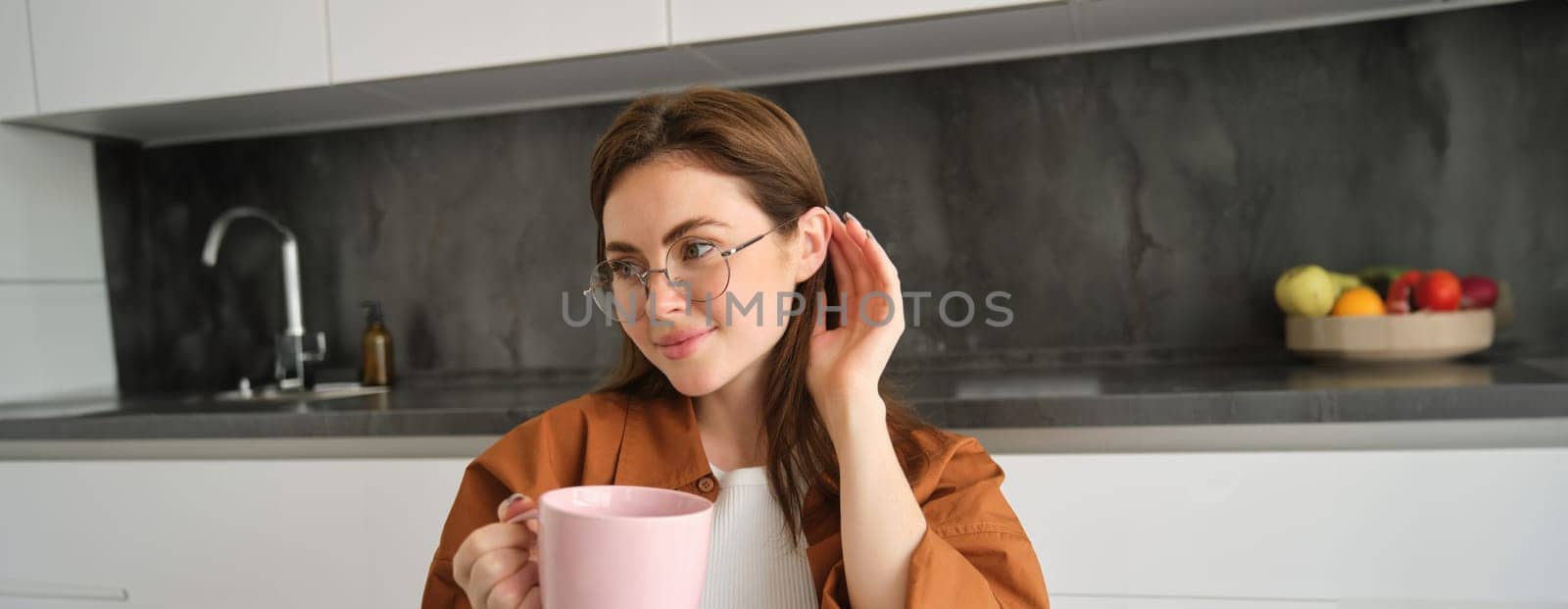 Portrait of lovely, tender young woman in glasses, sits at home with cup of tea. Girl in kitchen, enjoys drinking aromatic coffee by Benzoix