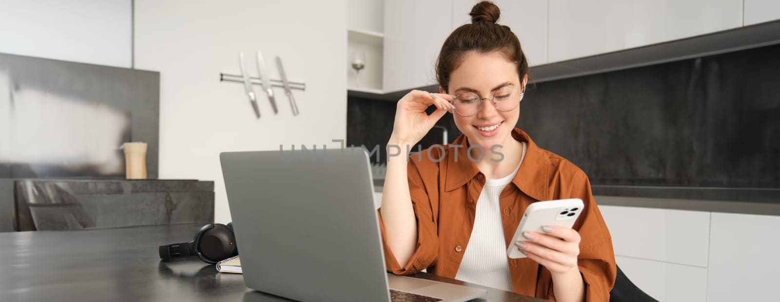 Portrait of young self-employed woman, replying to customers on smartphone, using laptop to check orders, sitting at home.