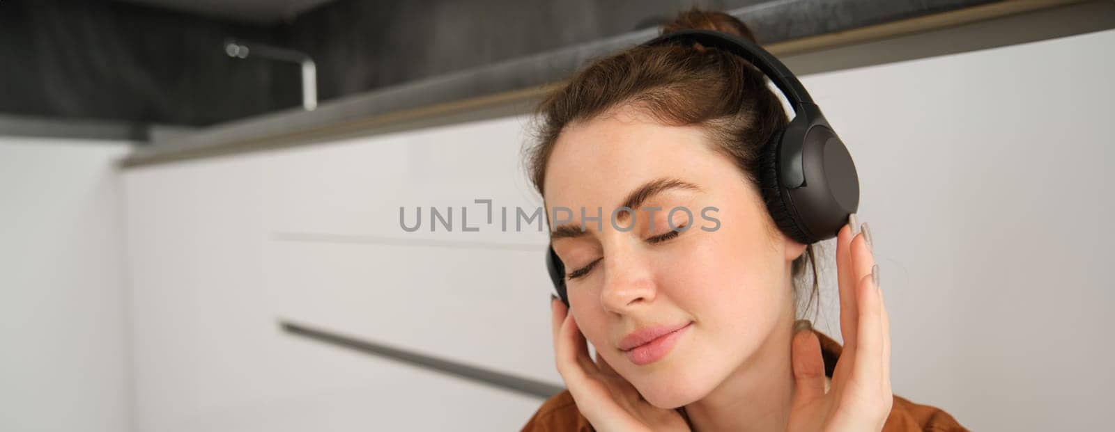 Portrait of attractive young woman in wireless headphones, smiling with closed eyes, relaxing with favourite songs on playlist.