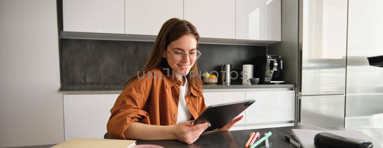 Portrait of young student, woman studying at home, working remotely, setup workplace in her kitchen, sitting on chair with digital tablet, reading in glasses.