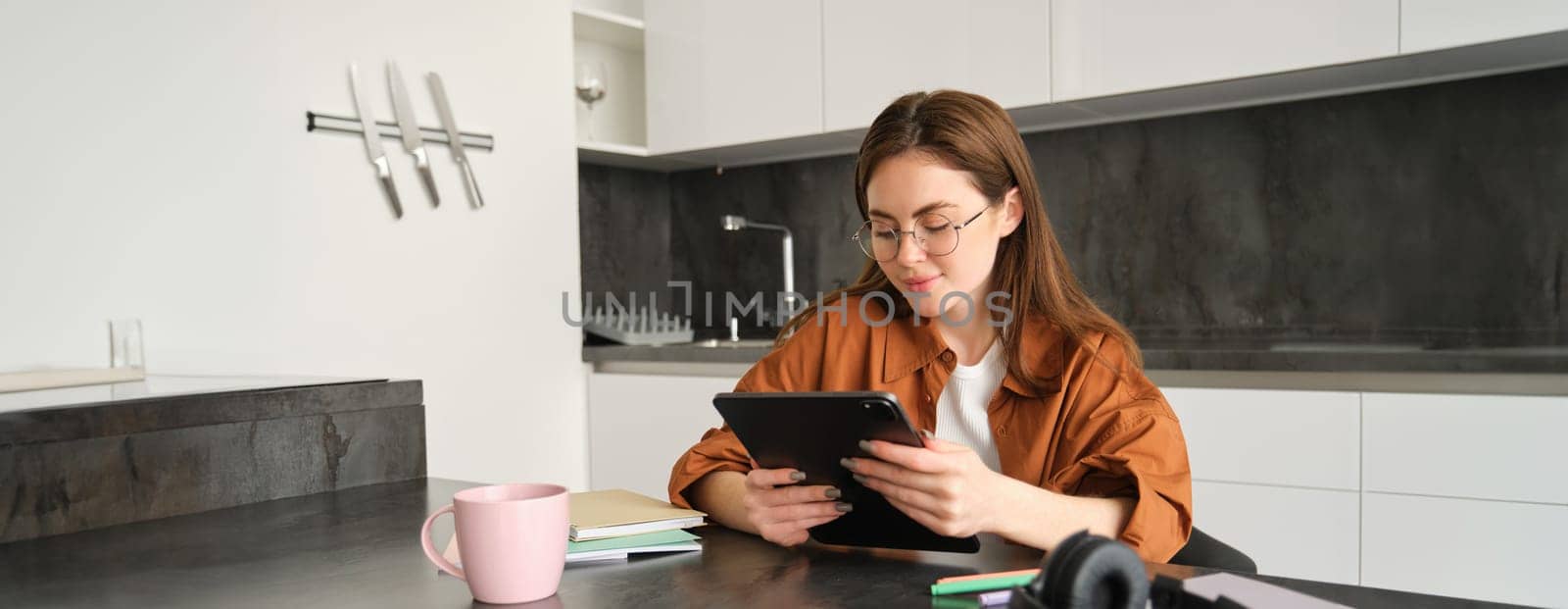 Portrait of beautiful young woman in glasses, reading on digital tablet, watching video, studying at home remotely, sitting in kitchen with headphones, papers and notebooks.