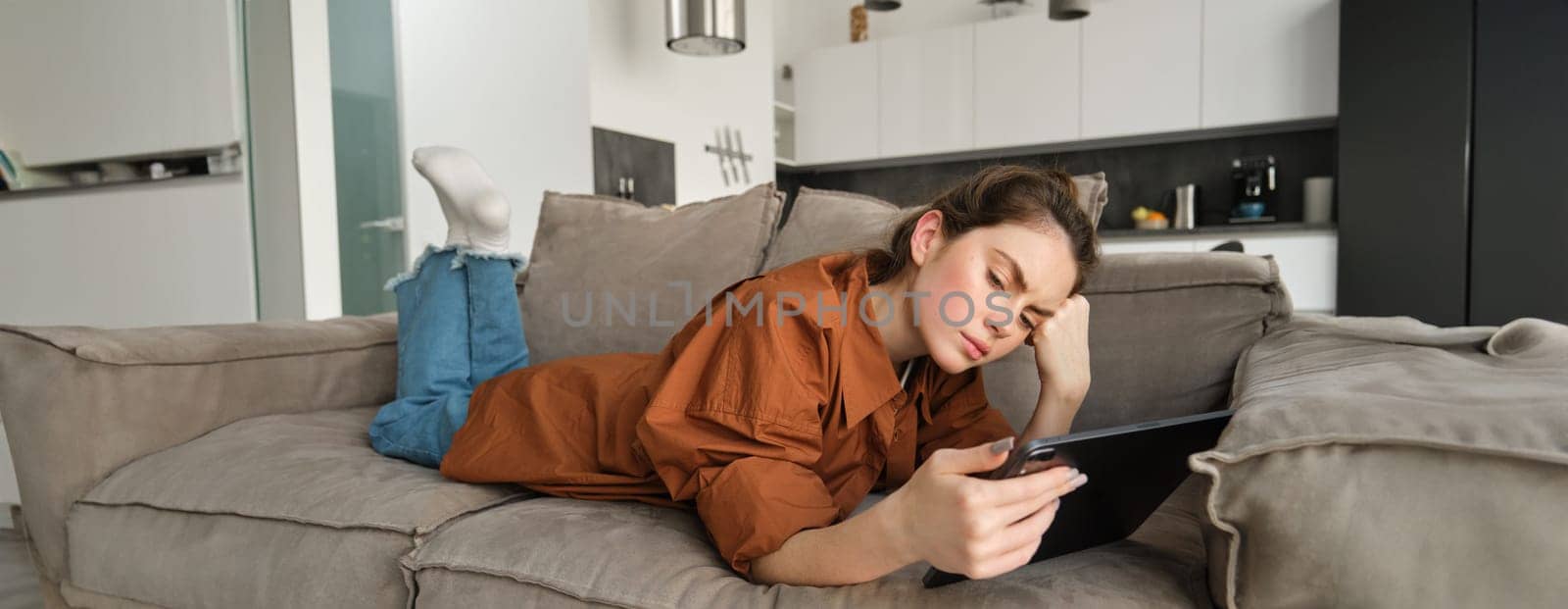 Portrait of woman with sad face, lying on couch at home, resting in living room with digital tablet, frowning and looking frustrated.