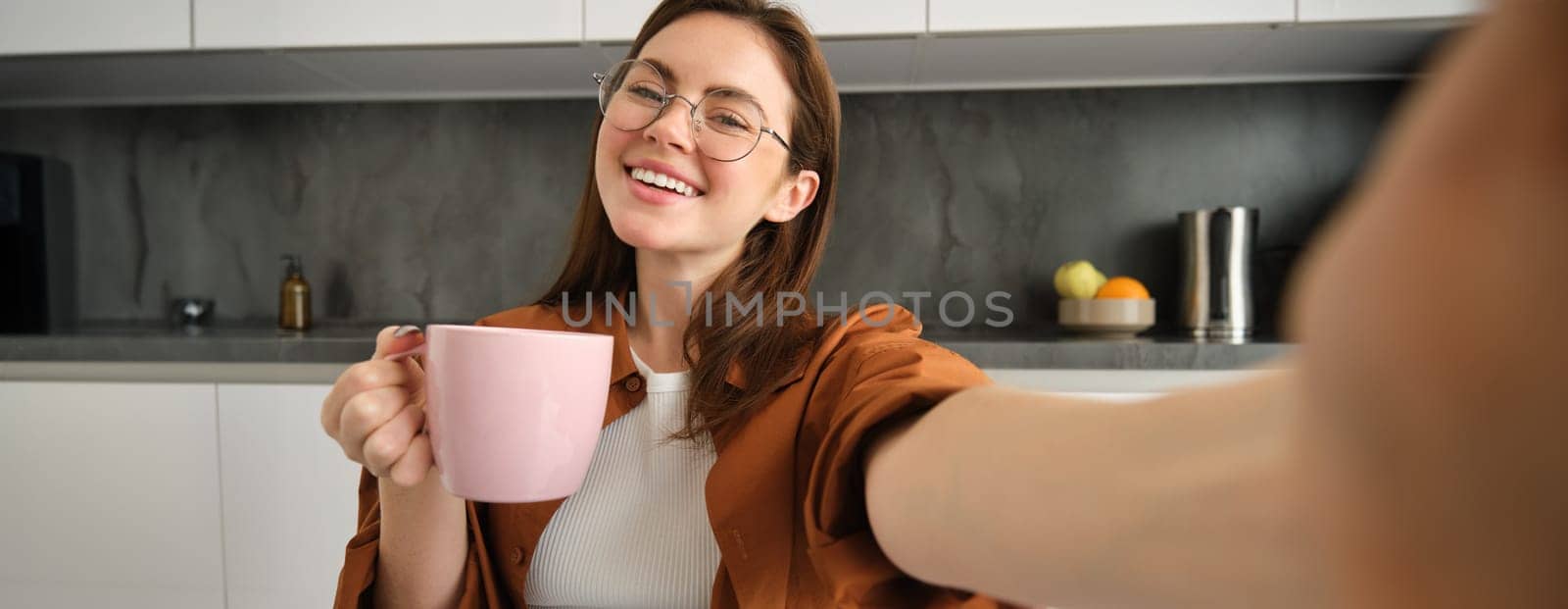 Carefree girl in glasses, takes selfie with smartphone, smiling and looking happy, enjoying hot drink in kitchen at home.