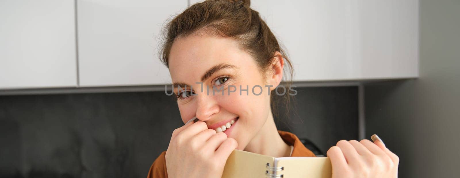 Beautiful modern woman, holding notebook near face, smiling and laughing, looking with flirting eyes at camera.