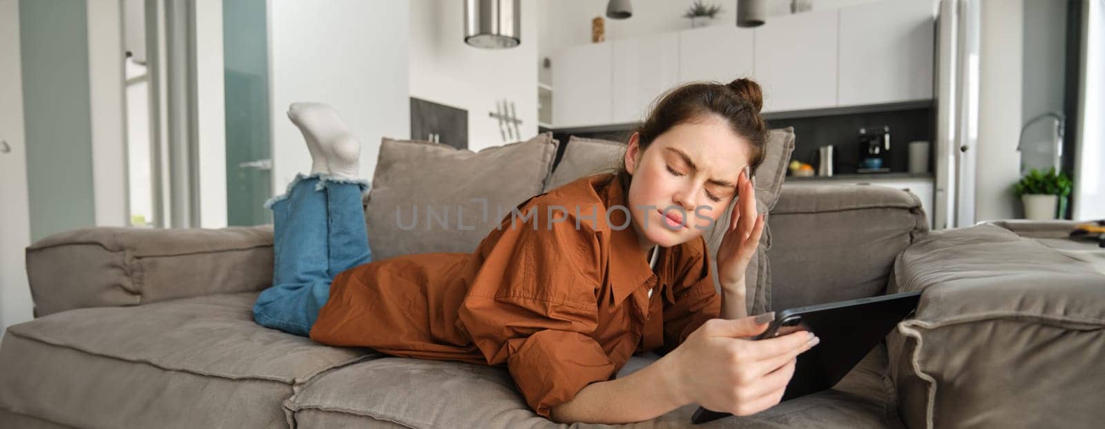 Portrait of young woman feeling unwell, lying on couch with digital tablet, touching head and frowning from painful migraine, has headache.