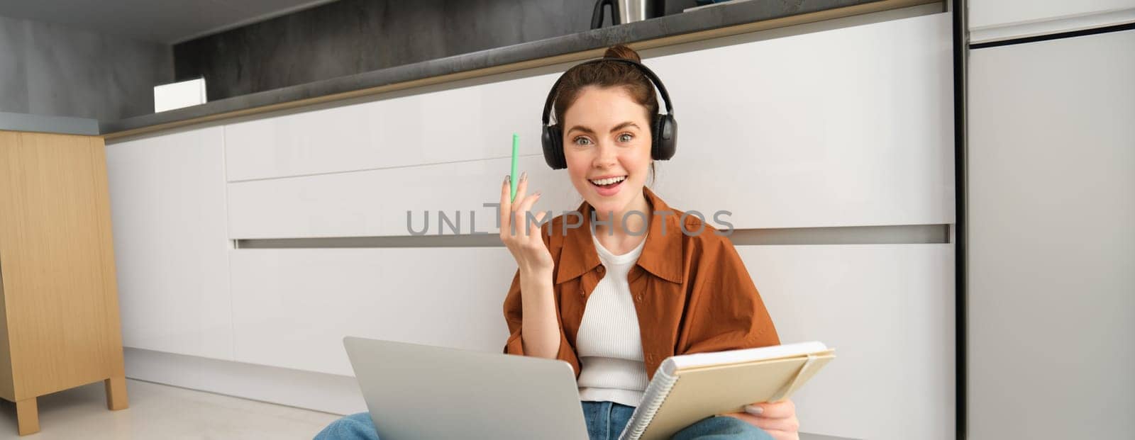 Portrait of young woman, modern student sits at home on floor and doing homework on laptop, listens music in headphones while making assignment or project for college by Benzoix