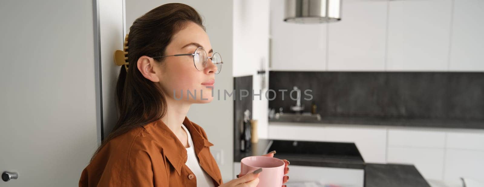 Portrait of cosy, beautiful and relaxed young woman at home, standing and leaning on wall with cup of coffee, drinking tea with thoughtful face expression by Benzoix